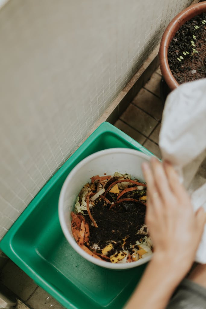 Photo by Letícia Alvares View of hands adding food scraps to a compost bin with visible soil.